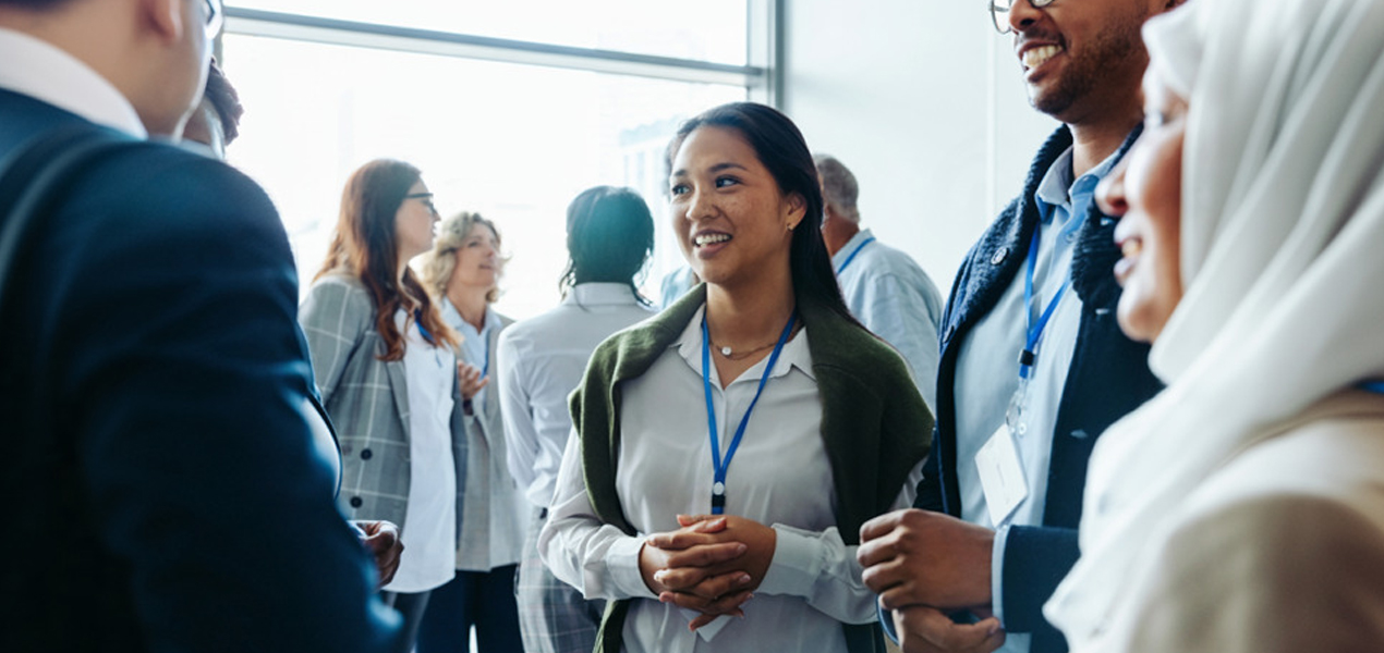 A group of people are mingling in a well-lit room, wearing business attire and name tags. A woman in the center has a sweater draped over her shoulders, smiling and engaging with others around her.